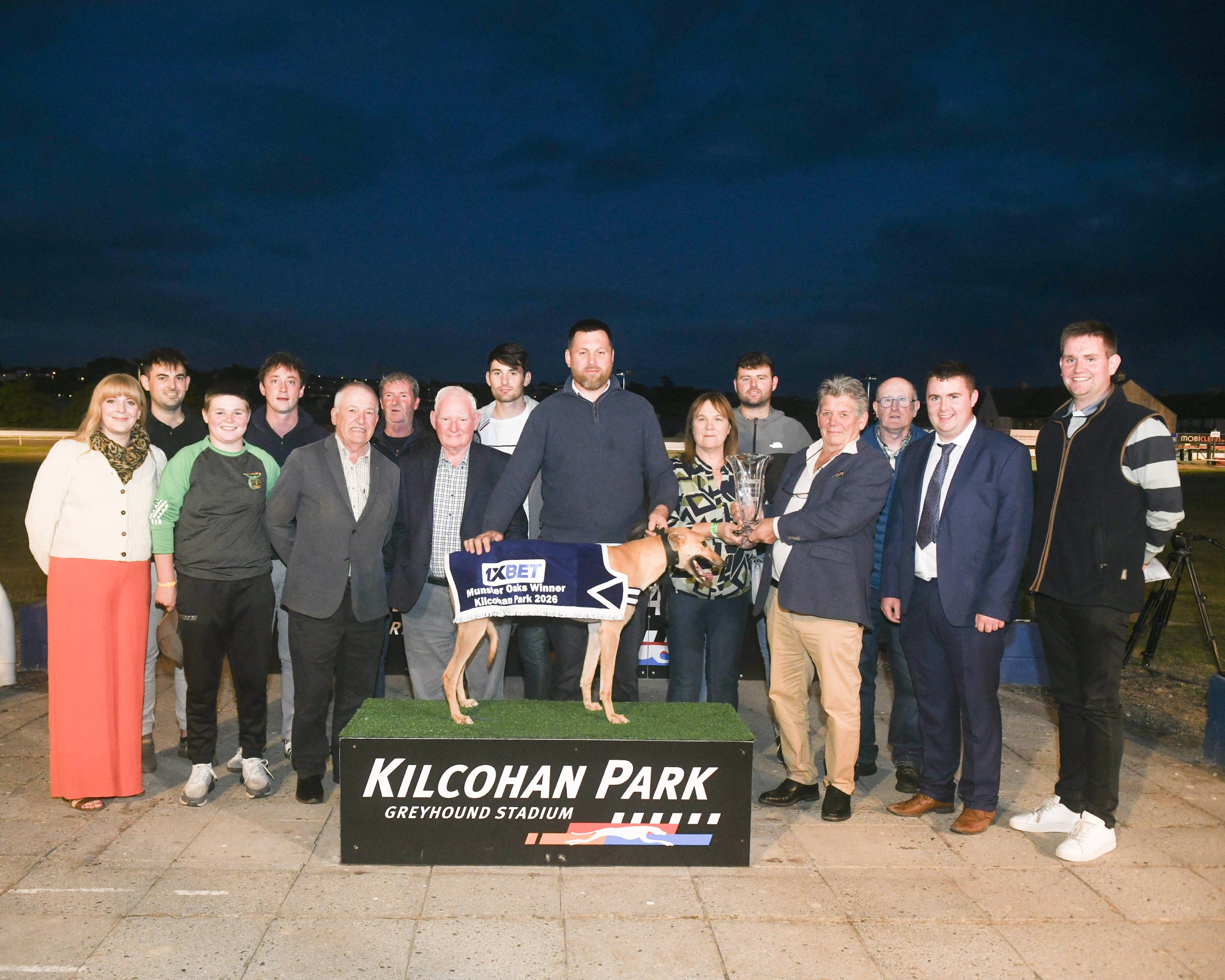 Image show a group presentation photograph at Kilcohan Park Greyhound Stadium showing the 1XBET Munster Oaks winner, Long Siesta, standing on a podium wearing a winner's jacket, surrounded by connections and race officials, with a trophy presentation taking place.