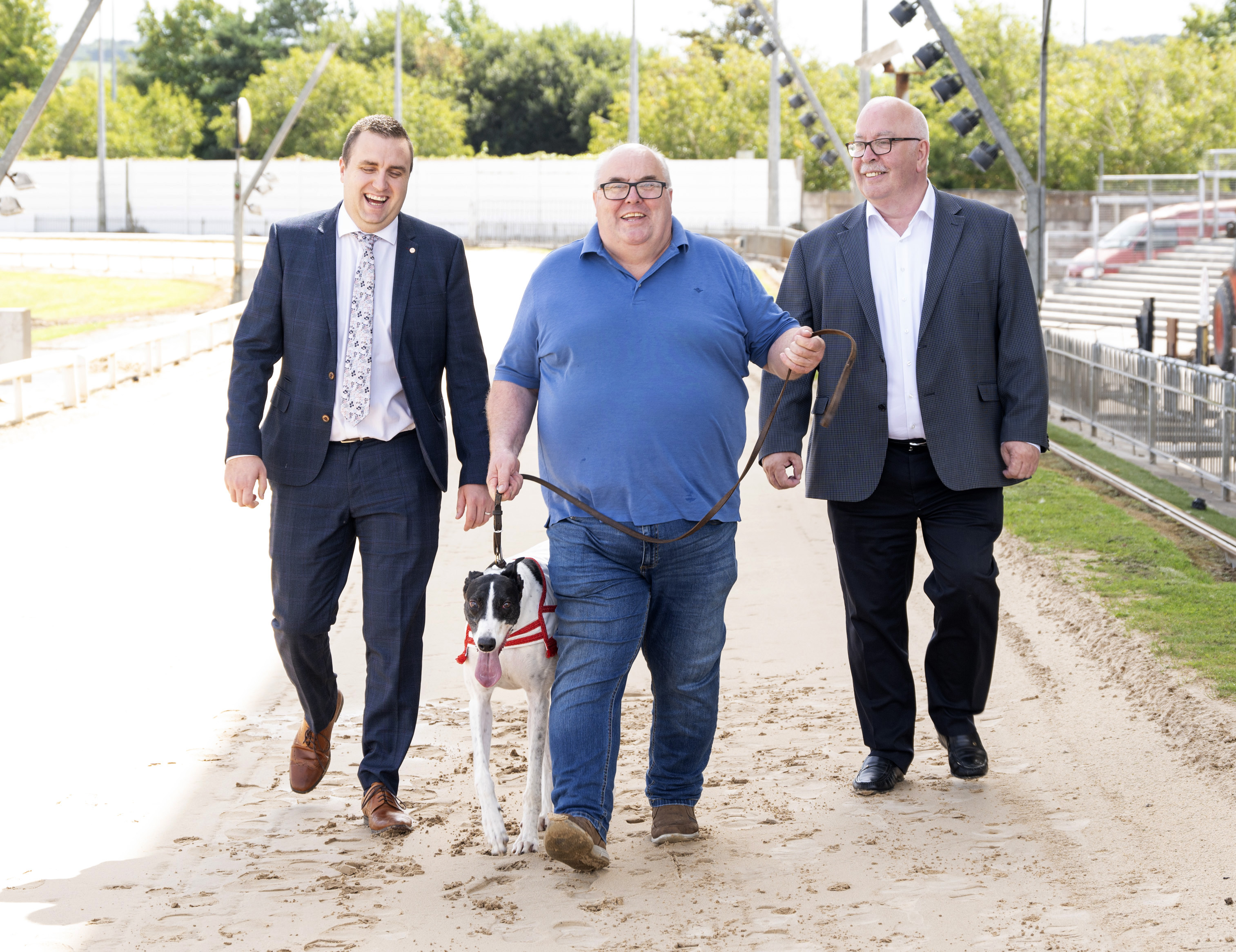 Image shows three men and a greyhound walking the track at Curraheen Park Greyhound Stadium at the launch of the 2024 Bar One Racing Irish Laurels